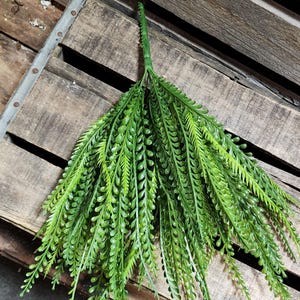 May include: A vibrant green artificial plant stem with long, slender leaves and a central stem. The leaves are a mix of light and dark green, creating a realistic appearance. The plant is displayed against a rustic wooden background.
