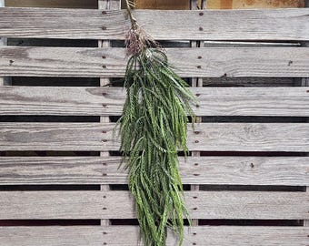 May include: A bundle of faux greenery hangs against a weathered wooden pallet backdrop. The greenery features long, slender, green leaves and a brown stem. The arrangement is tied with twine, creating a natural, rustic aesthetic.