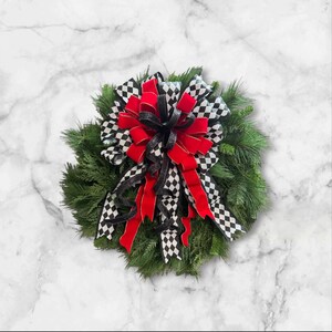 Puede incluir: Una corona decorativa con una base de agujas de pino verdes. La corona est&aacute; adornada con un gran lazo con cinta de terciopelo roja, cinta a cuadros en blanco y negro y cinta con purpurina negra. Un acento decorativo para el hogar.