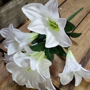 May include: A cluster of white artificial lilies with green stems and leaves, resting on a weathered wooden surface. The flowers have a trumpet shape with a green center.