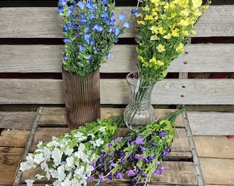 Puede incluir: Cuatro ramos de flores artificiales en diferentes colores: azul, amarillo, blanco y morado. Las flores están dispuestas en jarrones y en una caja de madera.