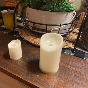 May include: Two white pillar candles with a warm glow, sitting on a wooden table. The candles are battery operated and have a realistic flame effect.