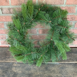May include: A circular evergreen wreath made of faux pine needles. The wreath is a vibrant green color and is displayed against a weathered wooden surface and a red brick wall. This wreath is suitable for seasonal home decor.