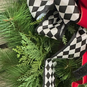 Puede incluir: Primer plano de una corona festiva con exuberantes ramas verdes de pino y cedro. Acentuada con un gran lazo de cinta a cuadros en blanco y negro y una cinta roja vibrante. Una pieza decorativa.