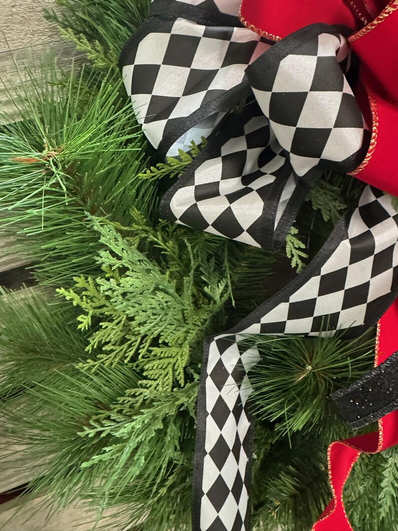 Puede incluir: Primer plano de una corona festiva con exuberantes ramas verdes de pino y cedro. Acentuada con un gran lazo de cinta a cuadros en blanco y negro y una cinta roja vibrante. Una pieza decorativa.
