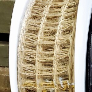 May include: A roll of natural jute netting, tightly woven in a grid pattern. The jute is a light brown color, and the roll is flanked by white plastic flanges. The netting is likely used for crafts or garden projects.