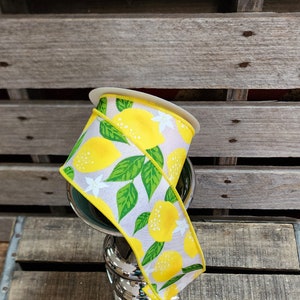 May include: A spool of lemon-themed ribbon with yellow lemons, green leaves, and white flowers on a white background. The ribbon is wrapped around a silver stand.