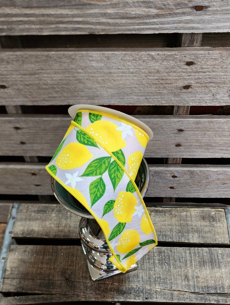 May include: A spool of lemon-themed ribbon with yellow lemons, green leaves, and white flowers on a white background. The ribbon is wrapped around a silver stand.