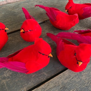 May include: Six red cardinal bird ornaments with pink feathers. The birds are made of felt and have black eyes and orange beaks.