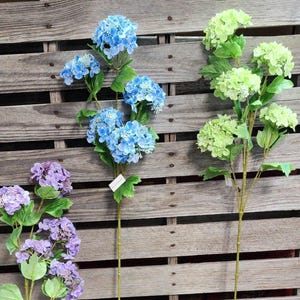 May include: Three stems of artificial hydrangea flowers in blue, green, and purple. The blue hydrangeas have multiple flower heads, while the green and purple hydrangeas have a single large flower head. The stems are green and the background is a wooden pallet.