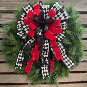 Puede incluir: Una corona festiva con un dise&ntilde;o llamativo. Presenta un gran lazo con un patr&oacute;n de cuadros blanco y negro, contrastado por cintas y acentos rojos vibrantes, todo sobre un fondo de follaje de pino verde.