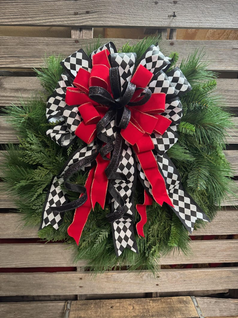 Puede incluir: Una corona festiva con un dise&ntilde;o llamativo. Presenta un gran lazo con un patr&oacute;n de cuadros blanco y negro, contrastado por cintas y acentos rojos vibrantes, todo sobre un fondo de follaje de pino verde.