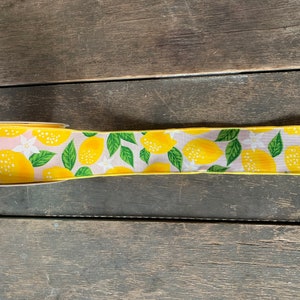 May include: A yellow ribbon with a lemon print. The ribbon has a light pink background with green leaves and white flowers.