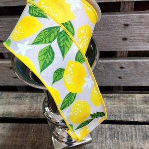 May include: A white ribbon with a yellow lemon print and green leaves. The ribbon is rolled up and sitting on a silver vase.