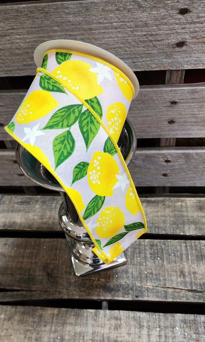 May include: A white ribbon with a yellow lemon print and green leaves. The ribbon is rolled up and sitting on a silver vase.