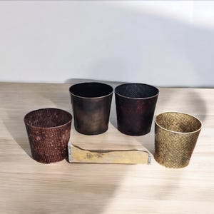 May include: Four metal planters in different colors and patterns. The planters are made of a textured metal and have a slightly distressed look. The planters are arranged in a row on a wooden surface.