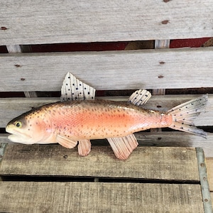 May include: A wooden sculpture of a rainbow trout, featuring a detailed design with pink, brown, and white hues. The fish has fins and a tail with black polka dots. The sculpture is displayed on a wooden surface.