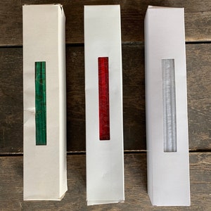 May include: Three rectangular white boxes with a vertical rectangular window revealing the contents. The boxes contain green, red, and silver items. The boxes are arranged on a weathered wooden surface.