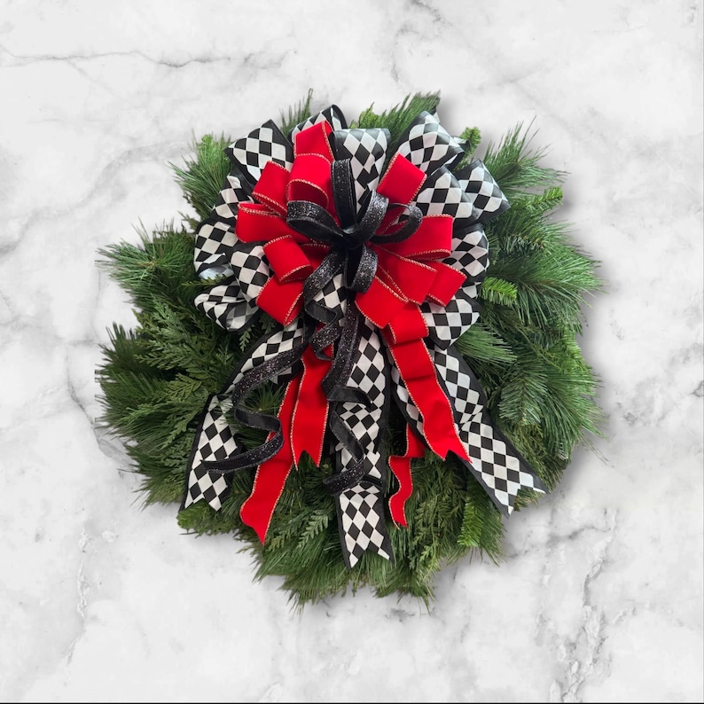 Puede incluir: Una corona redonda de hoja perenne con un gran lazo. El lazo presenta terciopelo rojo, purpurina negra y cinta a cuadros en blanco y negro. La corona se exhibe sobre un fondo de m&aacute;rmol blanco.