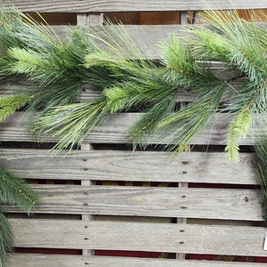 May include: Artificial pine garland with long, green needles and a mix of light and dark green hues. The garland is arranged on a weathered wooden pallet, creating a rustic and natural aesthetic. Ideal for holiday or seasonal decor.