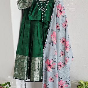 Pode incluir: Uma saia verde e prateada com uma blusa verde combinando e um lenço de estampa floral. A saia tem uma borda decorativa com bordados prateados. A blusa tem um fecho de botão de pérola. O lenço é de cor azul claro com flores cor-de-rosa.