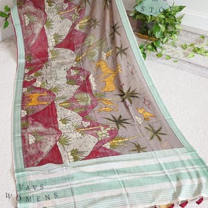 Op de afbeelding: Decoratief textiel met een gedetailleerde print met een landschap met figuren, bomen en dieren in grijstinten, rood en geel. De rand heeft een gestreept patroon in wit en lichtgroen, met rode kwastjes. De tekst "VAVS WOMENS STOP" is zichtbaar.