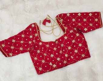 May include: A red blouse with gold floral embroidery and a square neckline. The long sleeves and neckline are trimmed with gold detailing. The blouse features a gold drawstring with a tassel.