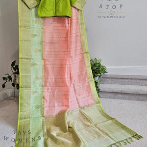 May include: A traditional Indian saree with a light green and peach color scheme. The saree features a detailed border and a matching green blouse. The text "VAVS WOMENS STOP" and "Six Yards Of Grandeur" is visible.