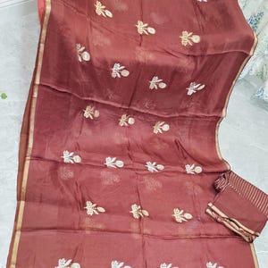 May include: A maroon-colored saree with gold trim and floral embroidery. The sheer fabric drapes elegantly, showcasing intricate detailing. A matching fabric piece is folded neatly on the side. The saree is a traditional garment.