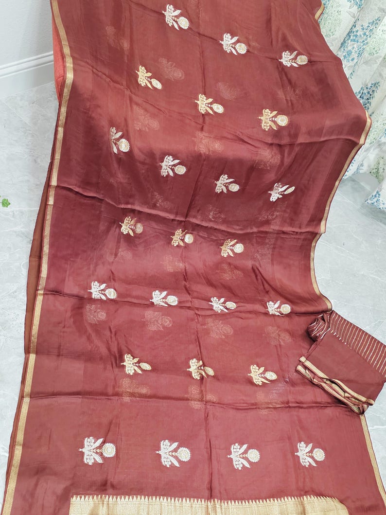 May include: A maroon-colored saree with gold trim and floral embroidery. The sheer fabric drapes elegantly, showcasing intricate detailing. A matching fabric piece is folded neatly on the side. The saree is a traditional garment.