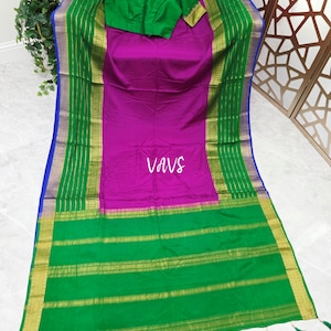 May include: A vibrant magenta saree featuring a wide green border with gold zari stripes and a matching green blouse with gold embroidery. The fabric displays the text VAVS in the center, and the bottom edge is finished with green tassels.