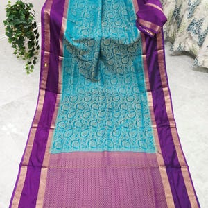 May include: A turquoise saree featuring a repeating paisley pattern in a lighter shade, bordered by wide purple bands with metallic gold trim. The bottom edge displays a dense geometric weave and purple tassels. Text reads VAVS WOMENS STOP.