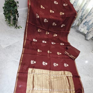 May include: A maroon-colored textile with gold floral embroidery and a gold border. The fabric is draped, showcasing the intricate design. A matching fabric piece is also visible.