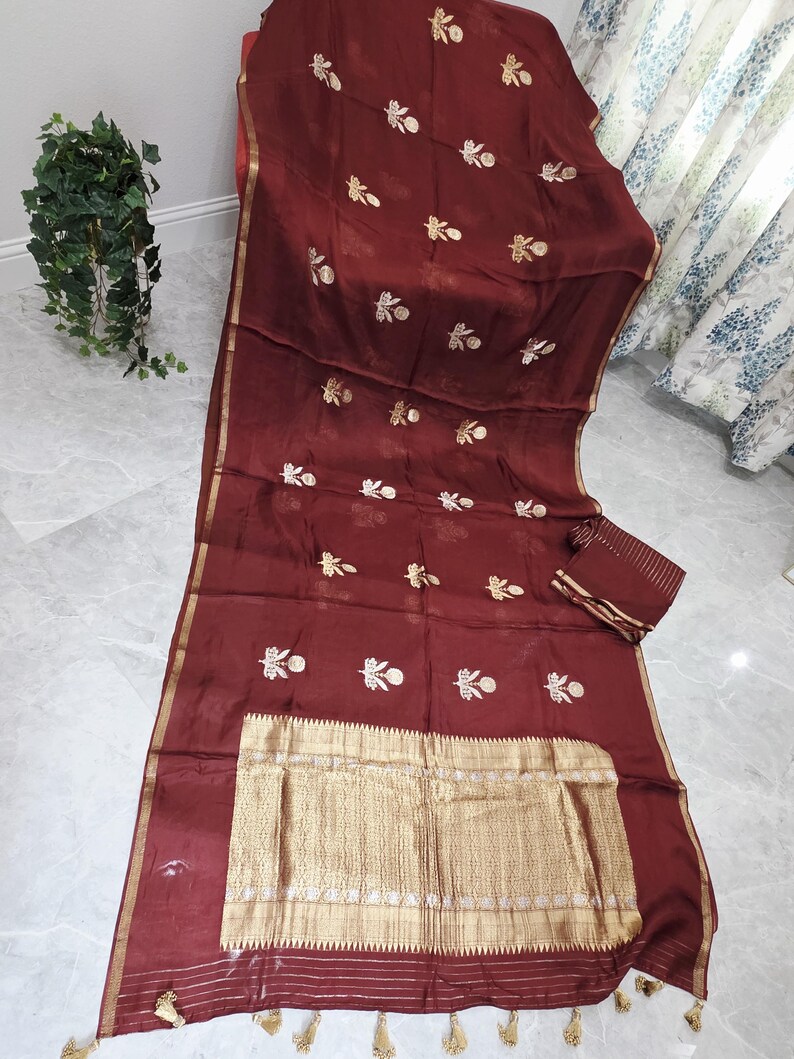 May include: A maroon-colored textile with gold floral embroidery and a gold border. The fabric is draped, showcasing the intricate design. A matching fabric piece is also visible.