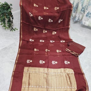 May include: A maroon textile with gold floral embroidery and a gold border. The fabric is draped, showcasing a matching small rectangular piece. Tassels adorn the bottom edge. The textile has a shiny appearance.