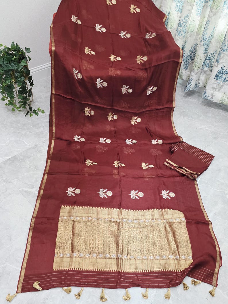 May include: A maroon textile with gold floral embroidery and a gold border. The fabric is draped, showcasing a matching small rectangular piece. Tassels adorn the bottom edge. The textile has a shiny appearance.