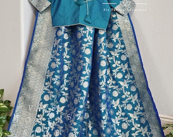 May include: A teal blue silk saree with a silver floral pattern and a matching teal blue blouse. The saree has a blue and silver border. The text "Six Yards of Grandeur" is visible in the image.