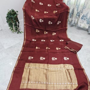 May include: A maroon textile with gold embroidery and trim. The fabric is draped over a chair, with a matching piece folded nearby. The gold trim features tassels at the edges. The embroidery is in a floral pattern.