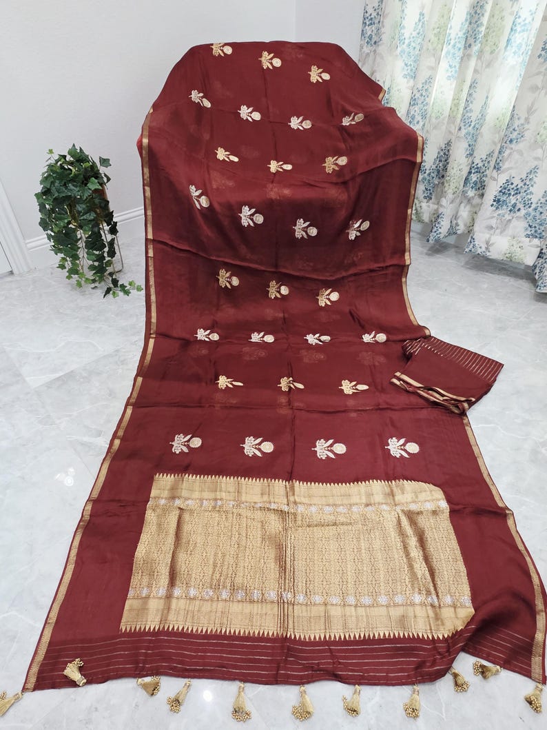 May include: A maroon textile with gold embroidery and trim. The fabric is draped over a chair, with a matching piece folded nearby. The gold trim features tassels at the edges. The embroidery is in a floral pattern.