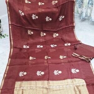 May include: A maroon-colored textile with gold and white floral embroidery. The fabric has a sheer quality, with a gold border and a matching folded piece. The design features repeating floral patterns.