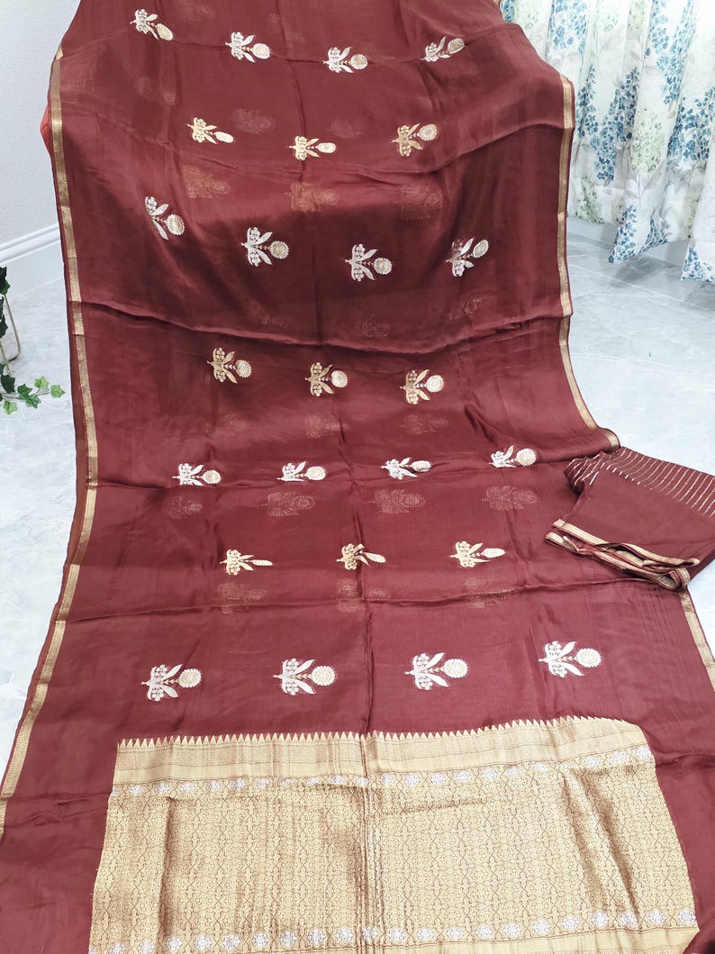 May include: A maroon-colored textile with gold and white floral embroidery. The fabric has a sheer quality, with a gold border and a matching folded piece. The design features repeating floral patterns.