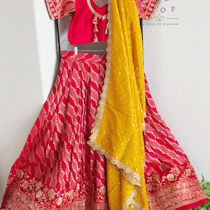May include: A traditional Indian outfit in red and gold, comprising a skirt, top, and scarf. The skirt has a detailed gold floral border and a repeating pattern. The top is a solid red with gold accents, and the yellow scarf has a gold lace trim.