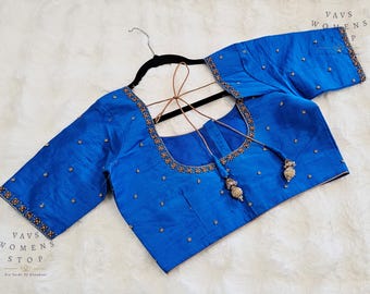 Op de afbeelding: Een helderblauwe blouse met ingewikkelde borduursels langs de halslijn en mouwranden. De blouse heeft delicate kralen en een vierkante halslijn. De mouwen zijn driekwart. De blouse hangt aan een zwarte hanger.