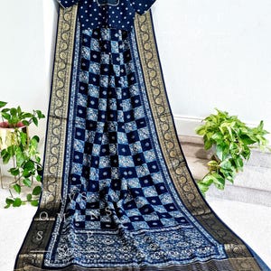 Op de afbeelding: Een zwart-blauwe saris met een gouden rand. De sari heeft een geruit patroon met bloemmotieven. De sari is over een hanger gehangen en heeft een bijpassende zwarte blouse met witte stippen.