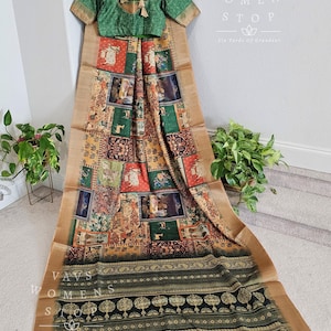 Op de afbeelding: Een groene blouse met een gouden kwast en een bedrukte zijden sari met een zwarte en gouden rand. De sari heeft een veelkleurige print met verschillende patronen en afbeeldingen. De tekst "Vavs Womens Stop Six Yards of Grandeur" is zichtbaar op de afbeelding.