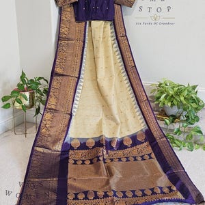 May include: A traditional cream and purple saree with a matching blouse. The saree features intricate gold patterns and a checkered design. The blouse is a deep purple with gold trim. The text "VAVS OMENS STOP Six Yards Of Grandeur" is visible.