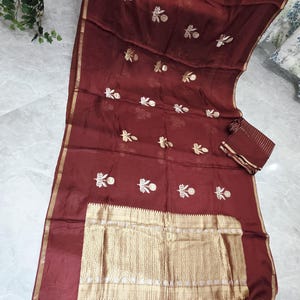 May include: A maroon-colored saree with gold embroidery and a gold border. The saree features floral embroidery and a rectangular gold panel with intricate detailing. A matching maroon fabric piece is included.