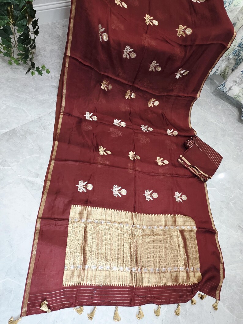 May include: A maroon-colored saree with gold embroidery and a gold border. The saree features floral embroidery and a rectangular gold panel with intricate detailing. A matching maroon fabric piece is included.