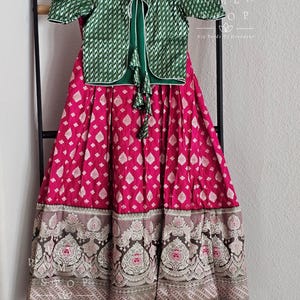 Könnte beinhalten: Ein grünes Oberteil mit weißem Muster und ein grüner Rock mit rosafarbenem und weißem Blumenmuster und einem silbernen und schwarzen Saum. Der Rock hat einen grünen Saum.