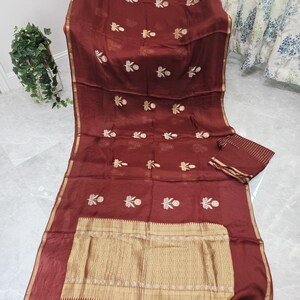 May include: A maroon-colored textile with gold embroidery and trim. The fabric features a repeating floral pattern and a decorative gold border. A matching piece of fabric is folded on the right side. Tassels hang from the bottom edge.